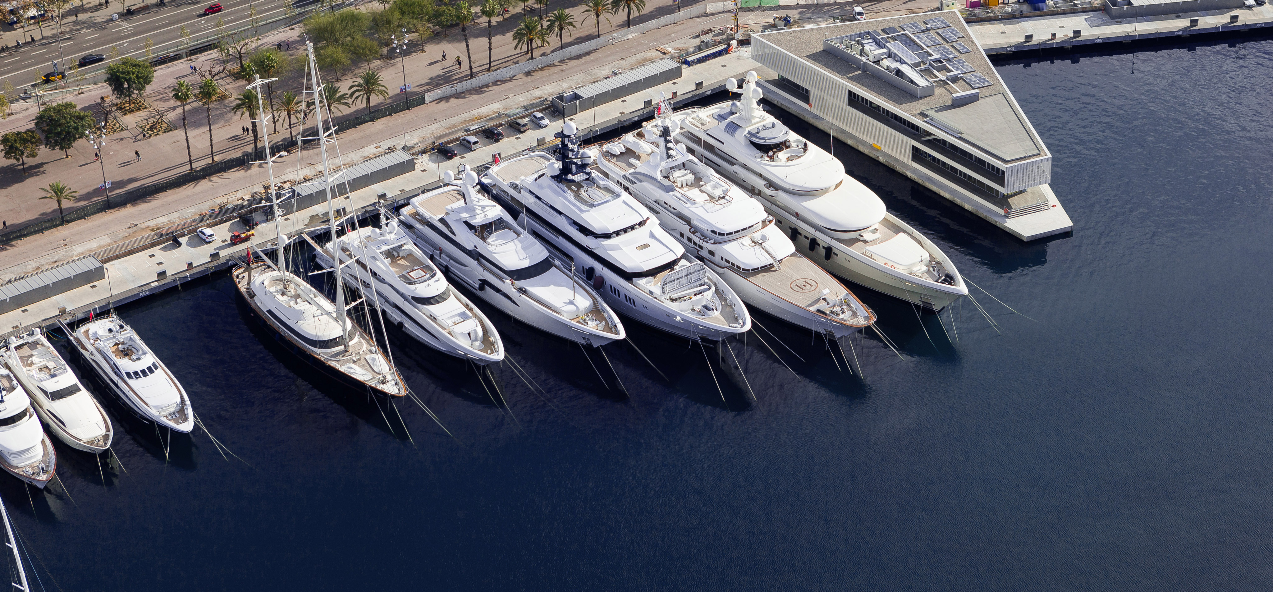 Multiple superyachts moored in a Barcelona marina with waterfront promenade and palm trees.